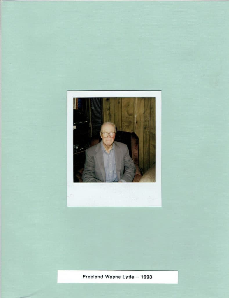 A Polaroid photo of an older man in glasses and a gray suit sitting in a chair, on a light green background with text "Freeland Wayne Lytle - 1993".