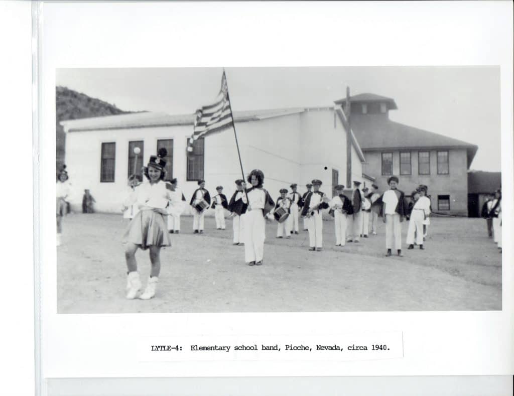 A black and white photo shows a young girl in a majorette uniform leading a school band with an American flag.