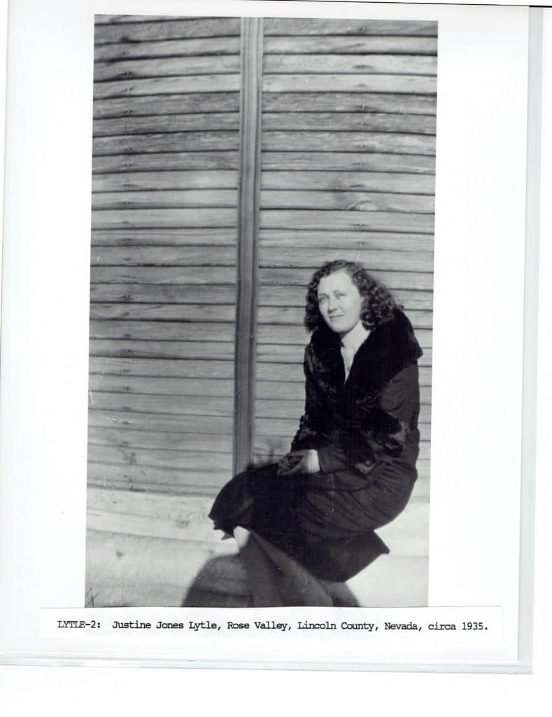 A black and white photo shows a woman in a dark coat, kneeling against a wooden wall.